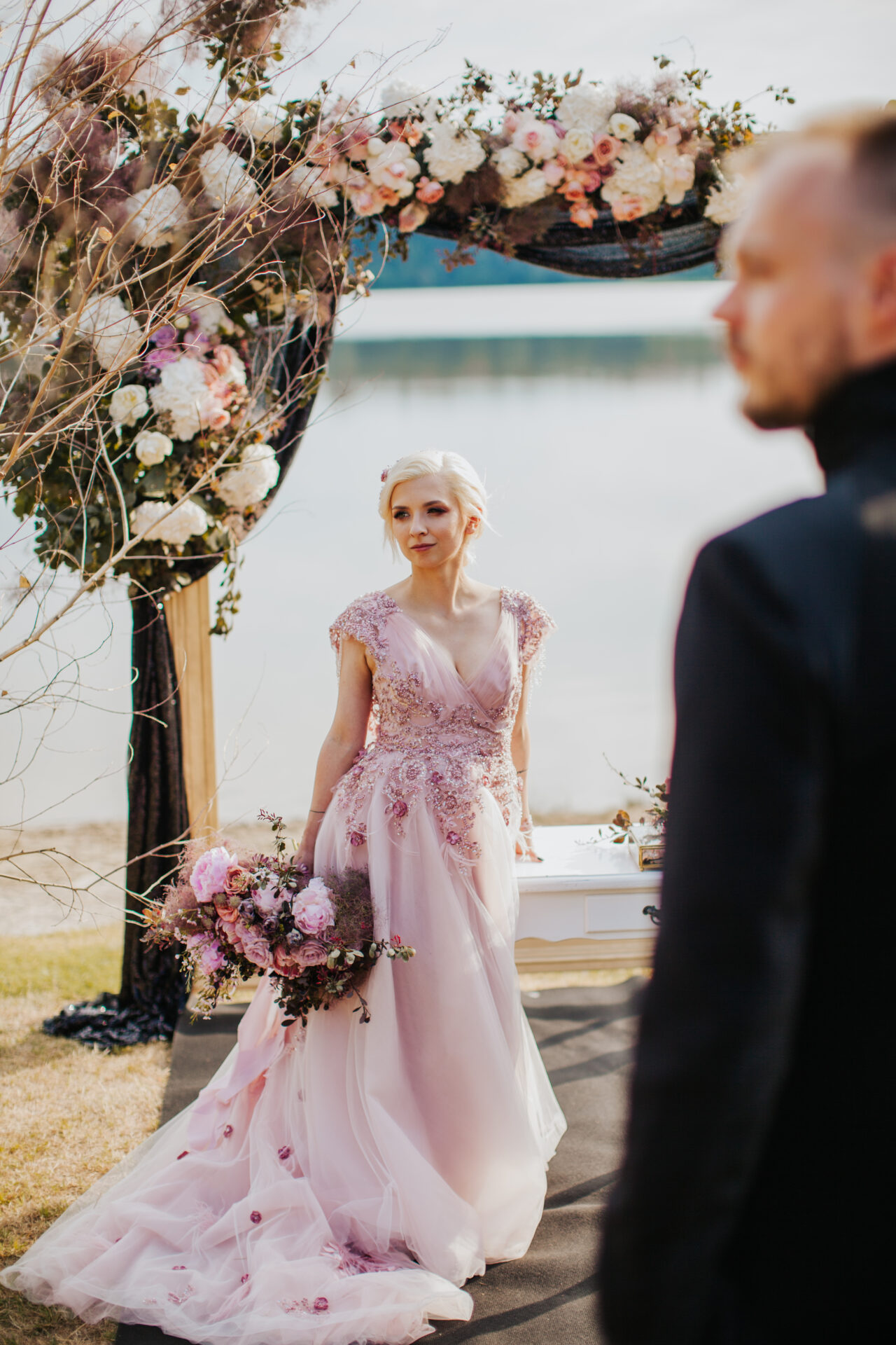 la novia con un lujoso vestido rosa se encuentra cerca de un arco de madera cubierto con material negro